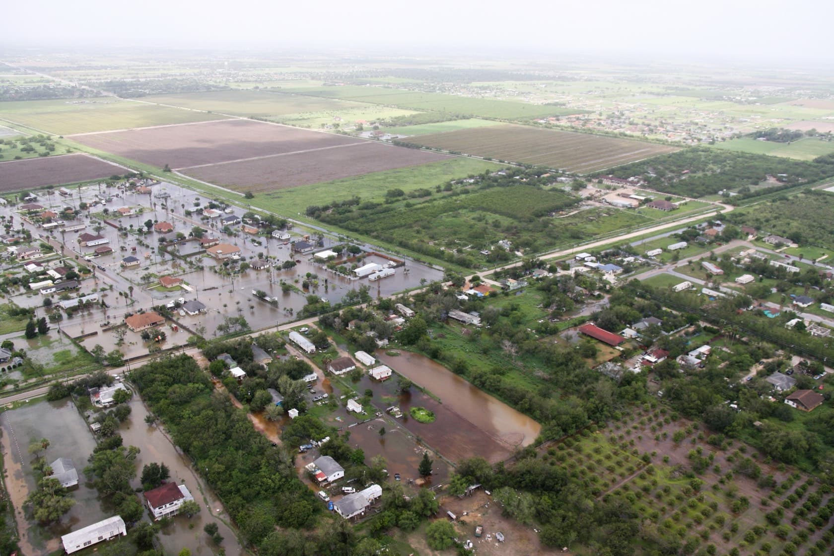 Aerial view of a flooded Texas neighborhood, streets and yards under brown floodwater while nearby blocks stay dry