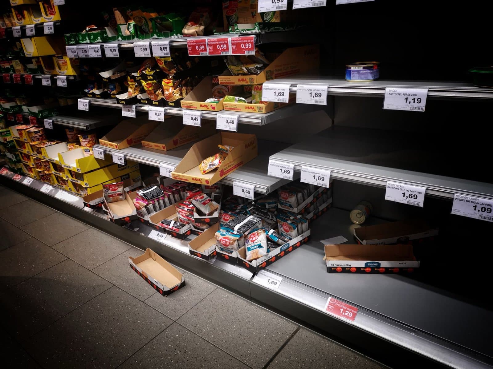 A dim supermarket aisle with half-empty shelves and scattered stock