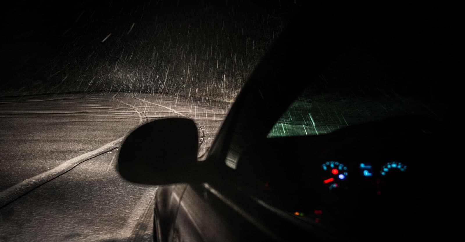 View from inside a car driving through heavy night snowfall, dashboard lights glowing