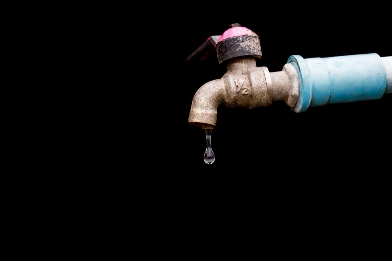 An outdoor faucet with a single drop of water falling, against a black background